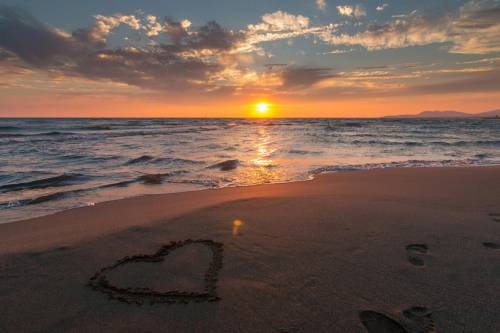 A heart shape drawn in the sand on a beach at sunset, with gentle waves in the foreground and a colourful sky with clouds.